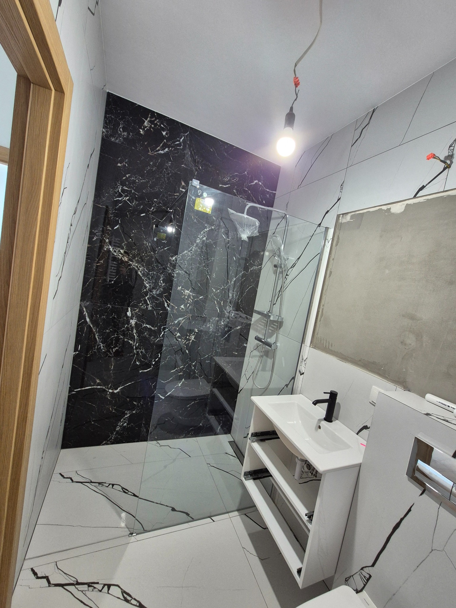 Nowoczesna łazienka z czarno-białymi płytkami imitującymi marmur, kabiną prysznicową typu walk-in i białą szafką pod umywalkę. Widoczna część ściany przed wykończeniem.