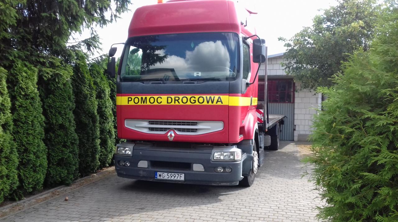 Czerwona ciężarówka z napisem 'POMOC DROGOWA' na żółtym pasie, zaparkowana na brukowanej drodze, otoczona zielonymi krzewami i drzewami.