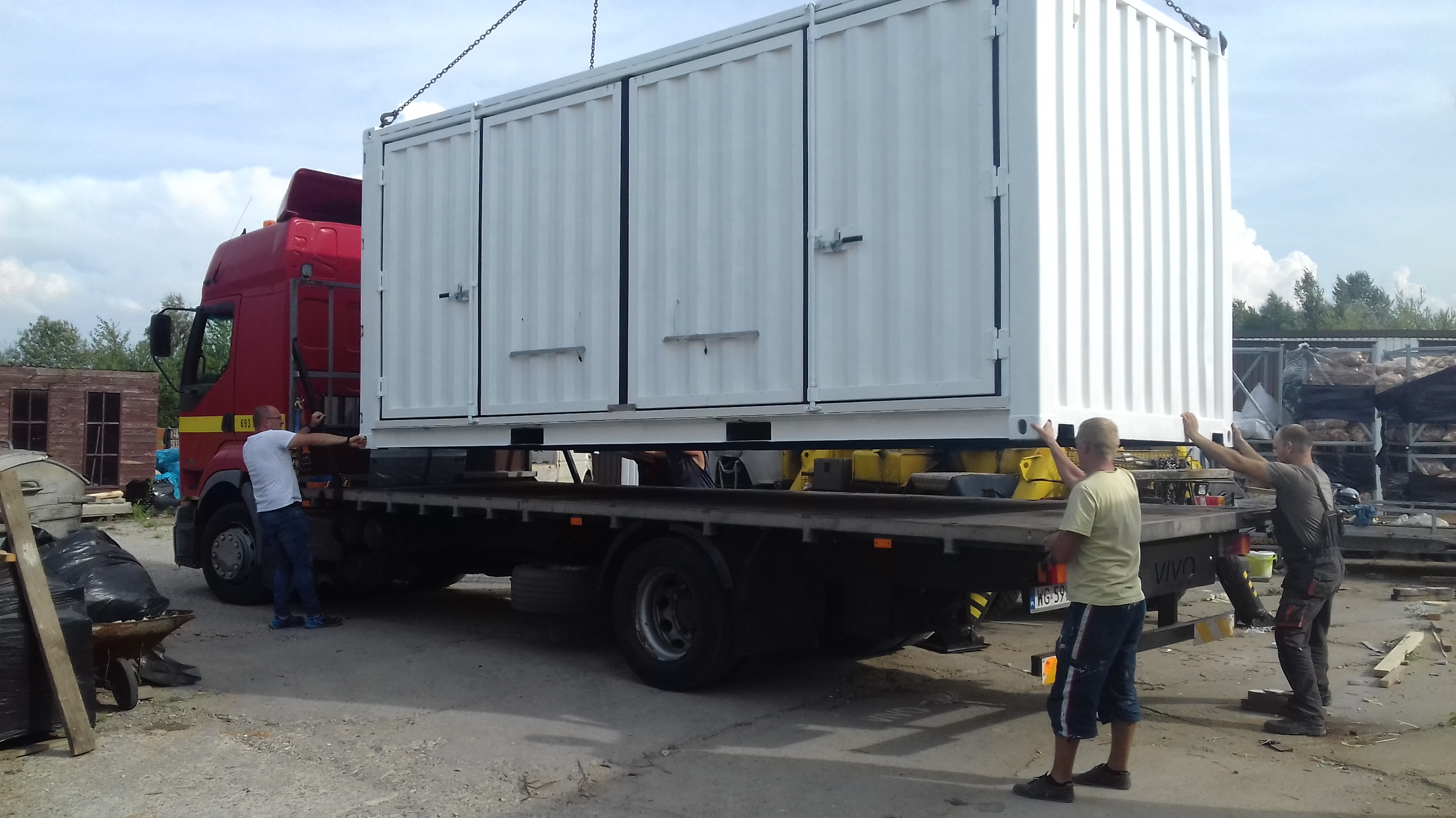 Ciężarówka z czerwoną kabiną przewozi biały kontener z dwuskrzydłowymi drzwiami, zabezpieczany przez trzech mężczyzn podczas załadunku.