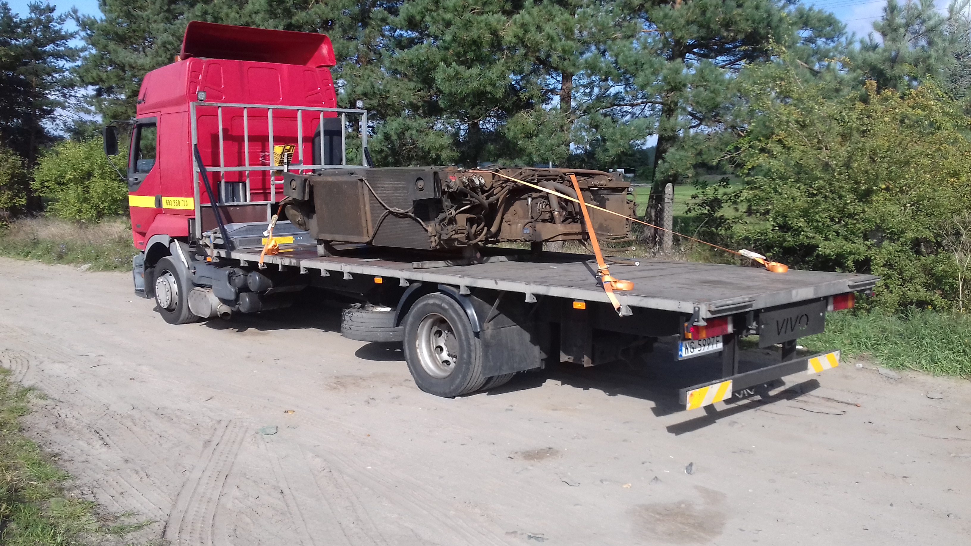 Czerwona ciężarówka z platformą transportową przewożąca duży, używany blok silnika zabezpieczony pomarańczowymi pasami, na tle drzew.