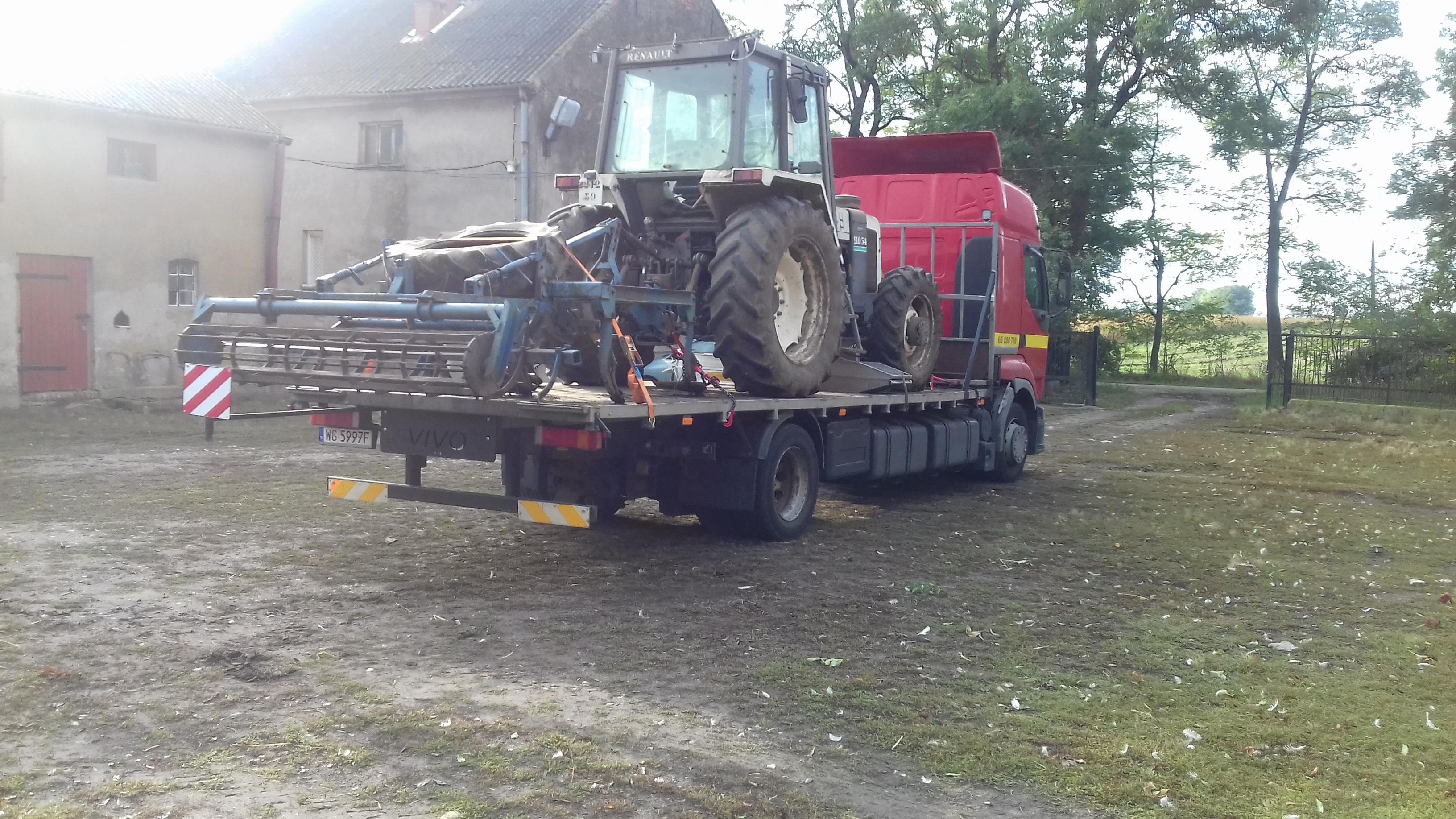 Czerwona ciężarówka z platformą przewozi traktor Renault i sprzęt rolniczy na tle budynku gospodarczego.