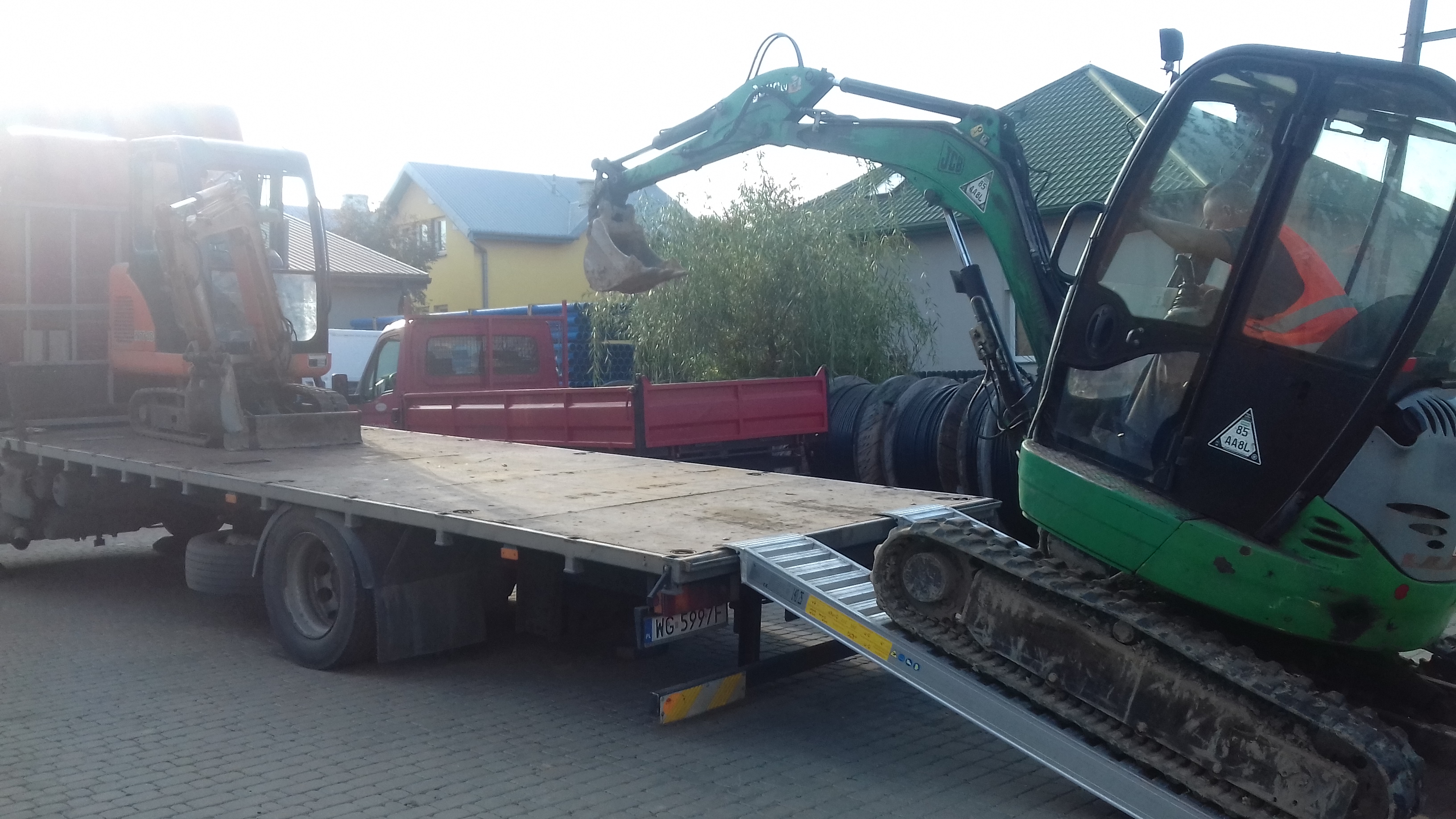 Załadunek zielonej mini koparki na platformę ciężarówki za pomocą rampy najazdowej; w tle widoczna druga, mniejsza koparka na platformie oraz budynki mieszkalne.