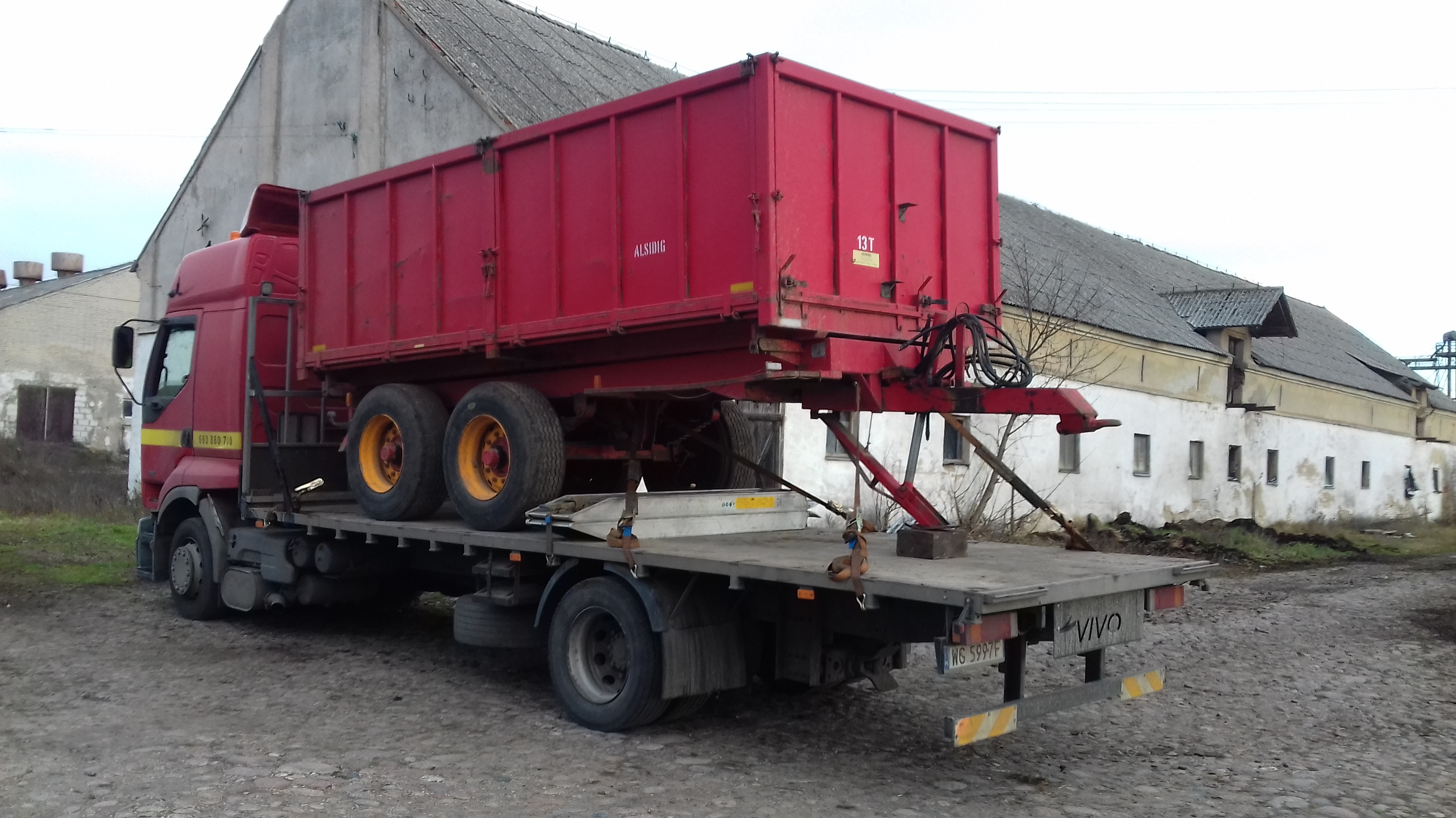 Czerwony zestaw transportowy: ciężarówka z platformą przewożąca czerwoną przyczepę kontenerową ALSIDIG, na tle zaniedbanych budynków gospodarczych.