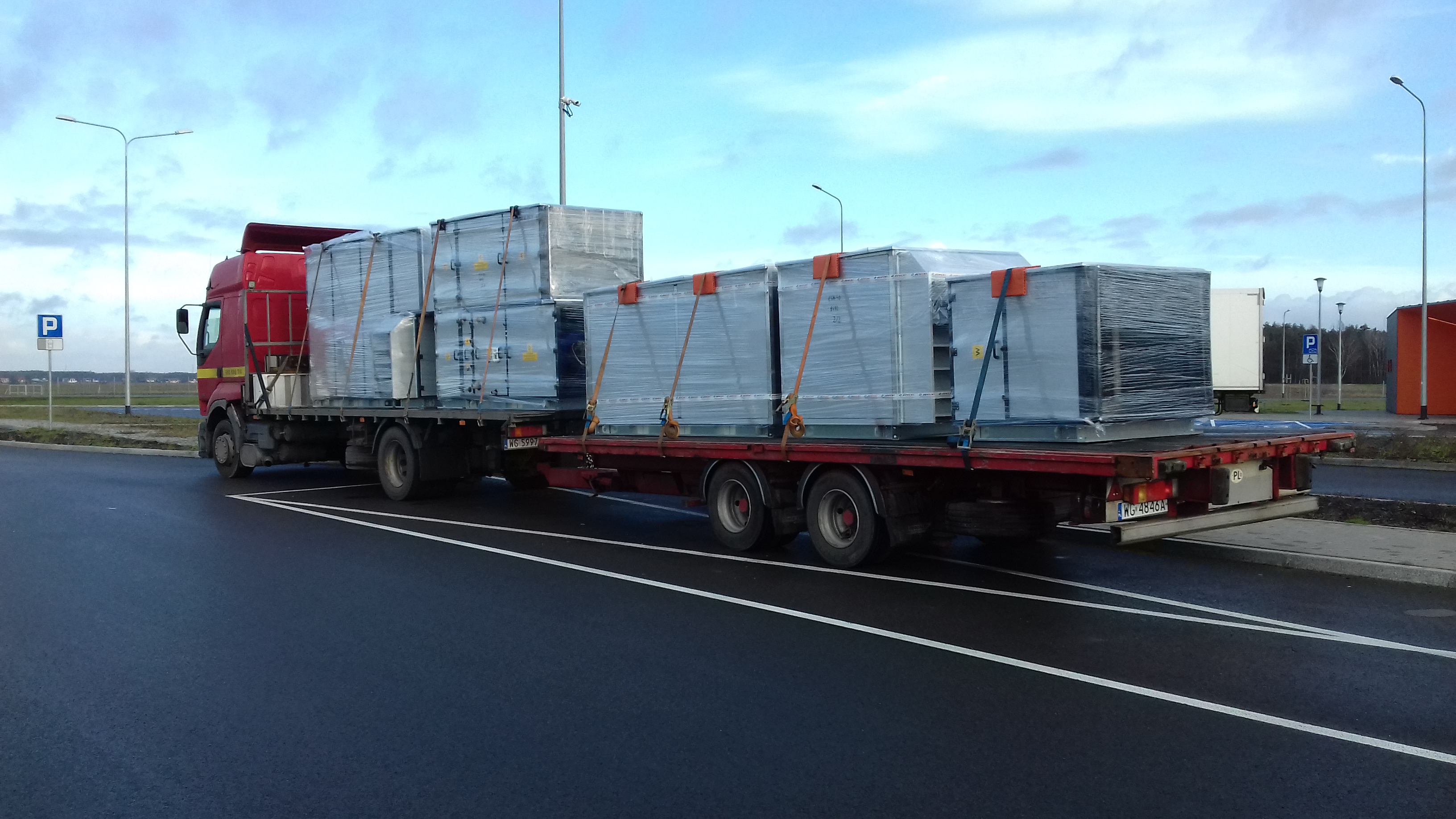 Czerwony samochód ciężarowy z naczepą przewozi kilka dużych, zapakowanych w folię kontenerów zabezpieczonych pasami transportowymi. Na pierwszym planie widoczna czarna, mokra nawierzchnia parkingu...