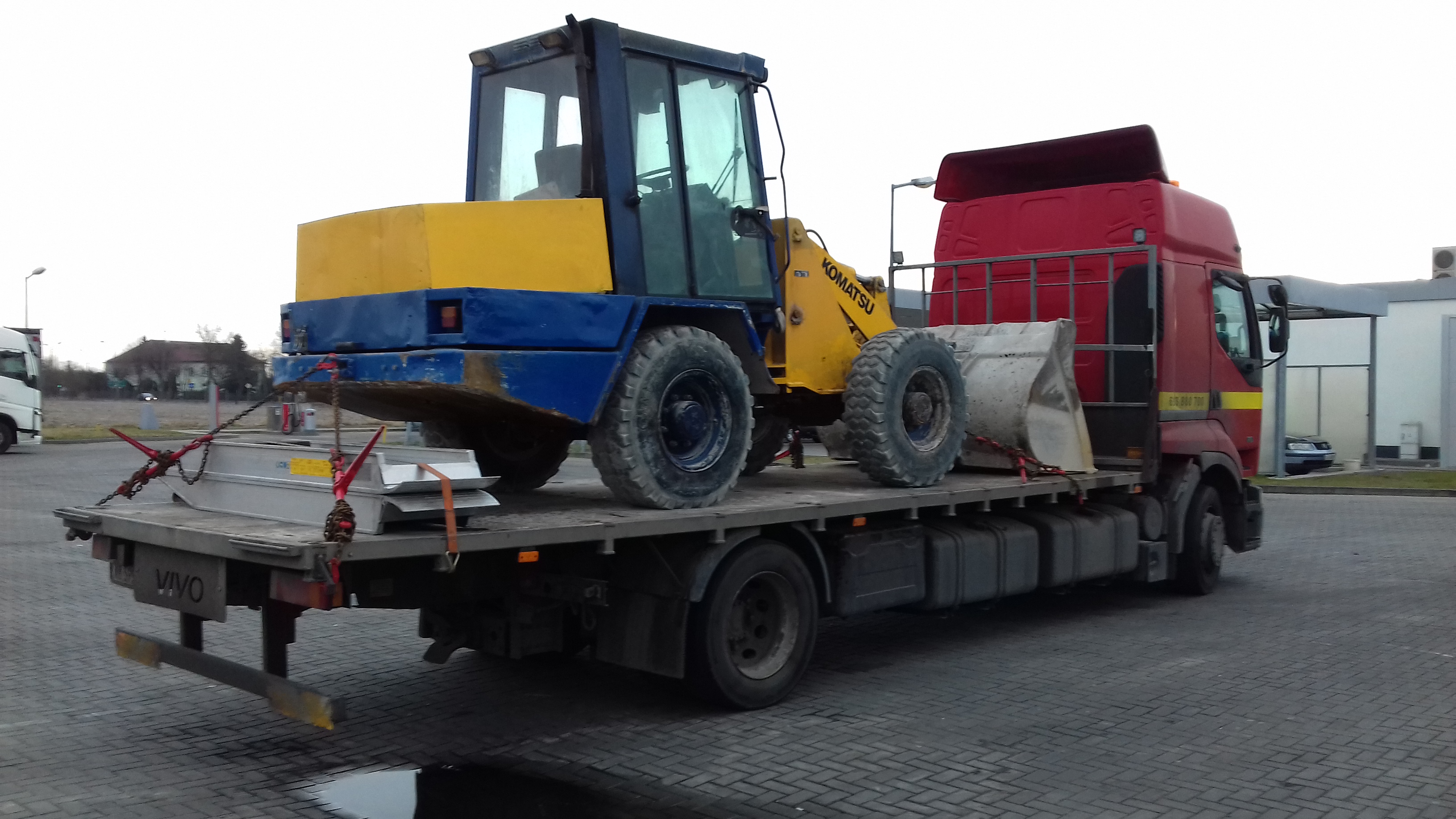 Żółto-niebieska ładowarka Komatsu zabezpieczona łańcuchami na platformie czerwonej ciężarówki VIVO, transportowana po brukowanym placu.