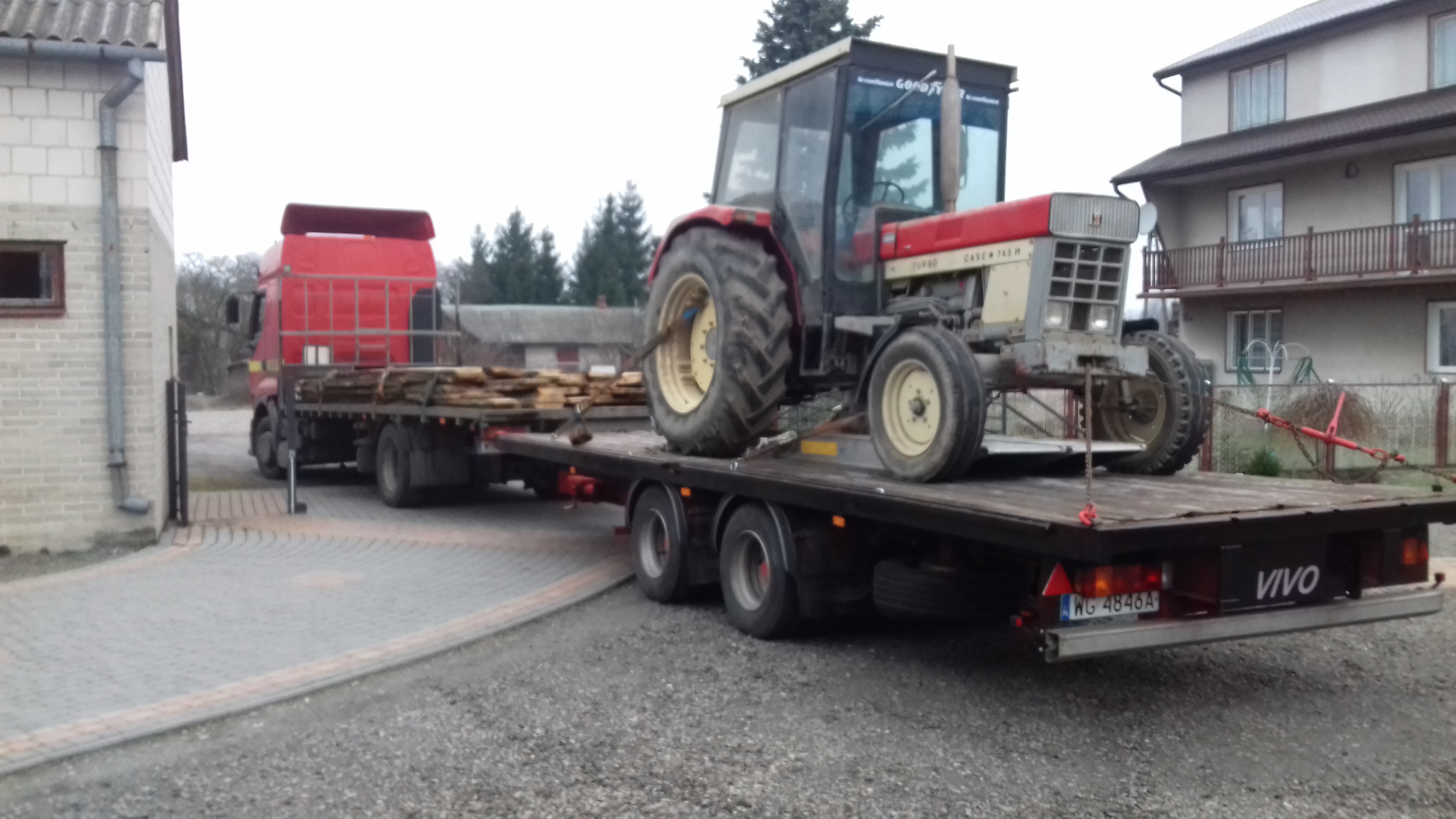 Czerwony ciągnik rolniczy Case 745 H z kabiną, przewożony na niskopodwoziowej przyczepie załadowanej drewnem, ciągniętej przez czerwony samochód ciężarowy