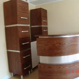 Recepcja z ciemnego drewna z srebrnymi akcentami, obok wysokie szafki z szufladami, widoczny detal usłojenia drewna i metalowe uchwyty.