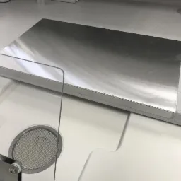 Metalowy blat roboczy na białej szafce z widocznym fragmentem przezroczystej osłony i kratki wentylacyjnej w tle blatu o szarej, nakrapianej powierzchni.