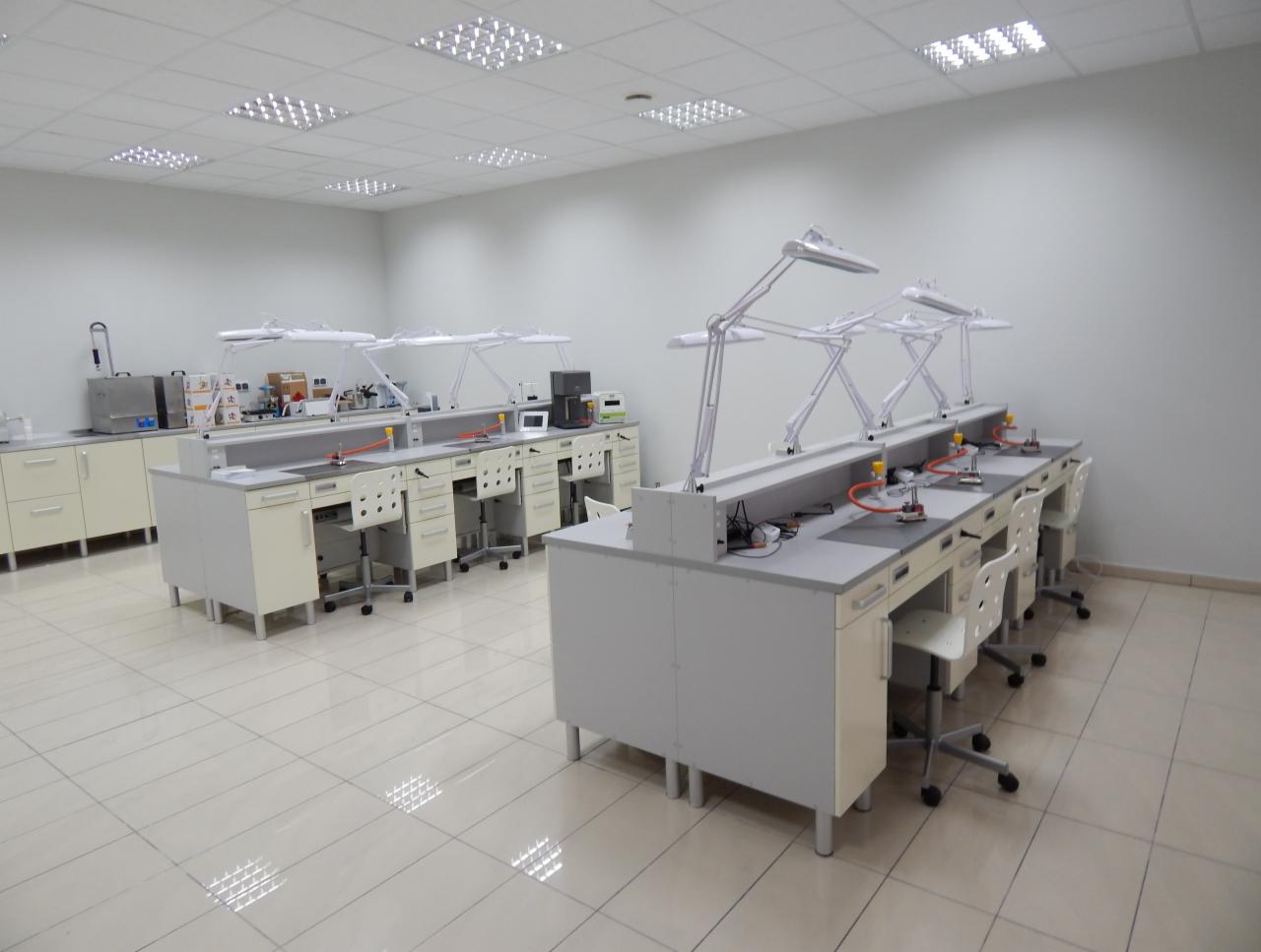 Jasne, przestronne laboratorium protetyczne z rzędami białych stanowisk roboczych wyposażonych w lampy i narzędzia, widok na sterylną salę z białymi ścianami i podłogą z płytek.