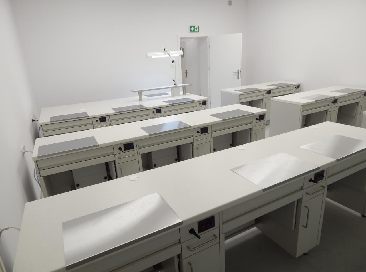 Wnętrze pracowni z rzędem białych, specjalistycznych stołów warsztatowych z szufladami i metalowymi nakładkami, oświetlonych lampami, z widocznym znakiem ewakuacyjnym nad otwartymi drzwiami.