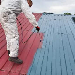 Pracownik w kombinezonie maluje blaszany dach na szaro za pomocą pistoletu natryskowego, widoczna część dachu pomalowana na czerwono, w tle komin i zieleń.