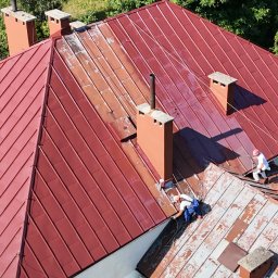 Alpin-Sky Specjalistyczne Usługi Wysokościowo- Budowlane Marcin Walewski - Częściowo umyty, czerwony dach z blachy trapezowej podczas pracy dwóch alpinistów na linach, widok z góry na dom z ceglanymi kominami i zielenią w tle.