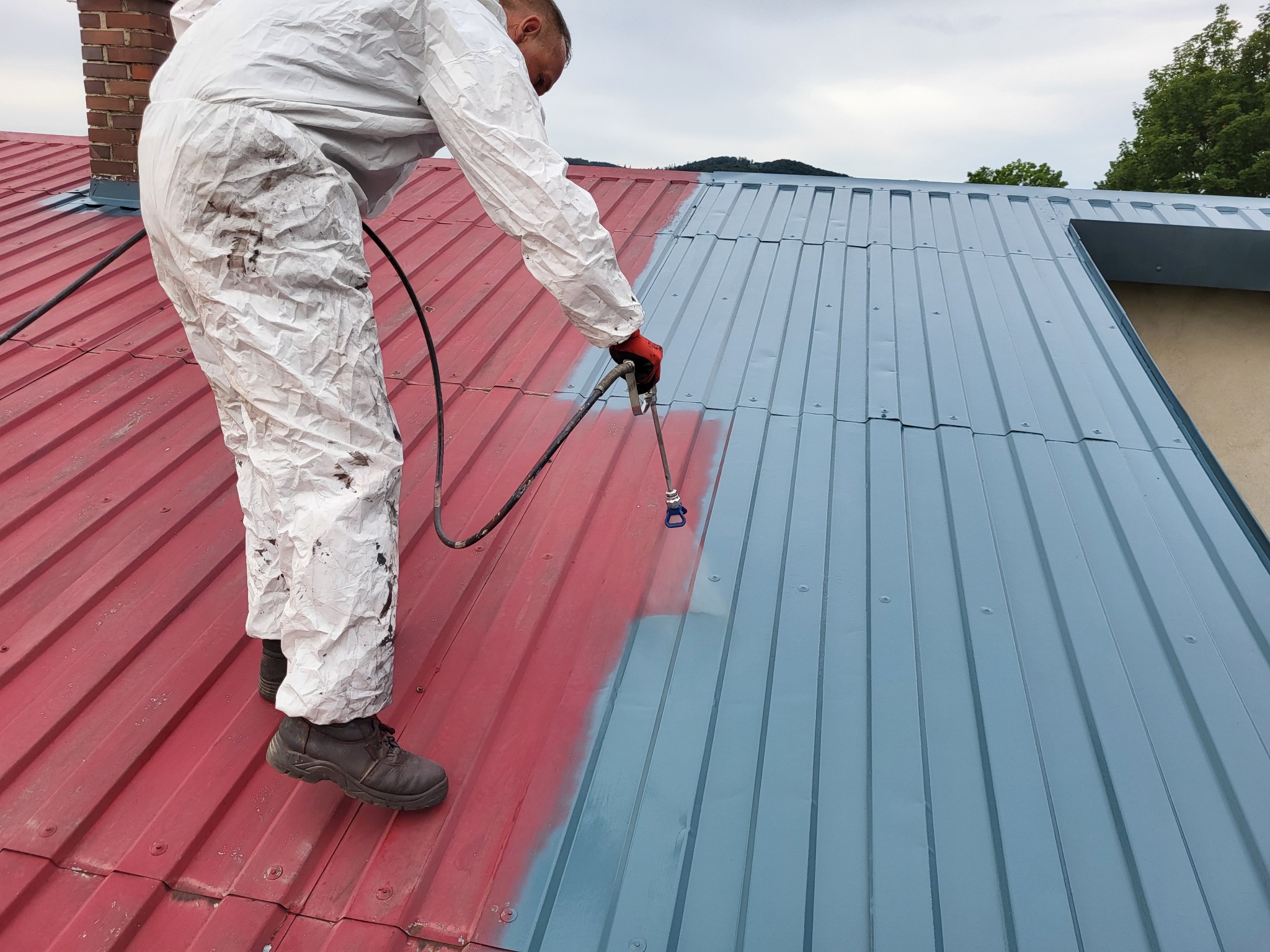 Pracownik w kombinezonie maluje blaszany dach na szaro za pomocą pistoletu natryskowego, widoczna część dachu pomalowana na czerwono, w tle komin i zieleń.