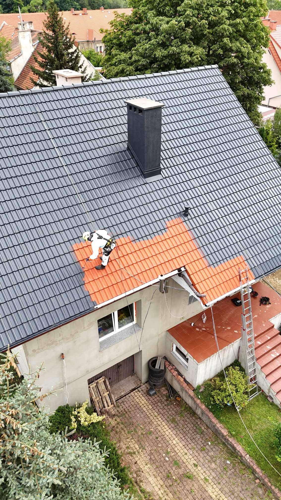 Pracownik w kombinezonie maluje dach z metalowej dachówki na pomarańczowo, kontrastując z szarym kolorem reszty dachu i ciemnym kominem, widok z góry na dom z podwórkiem.