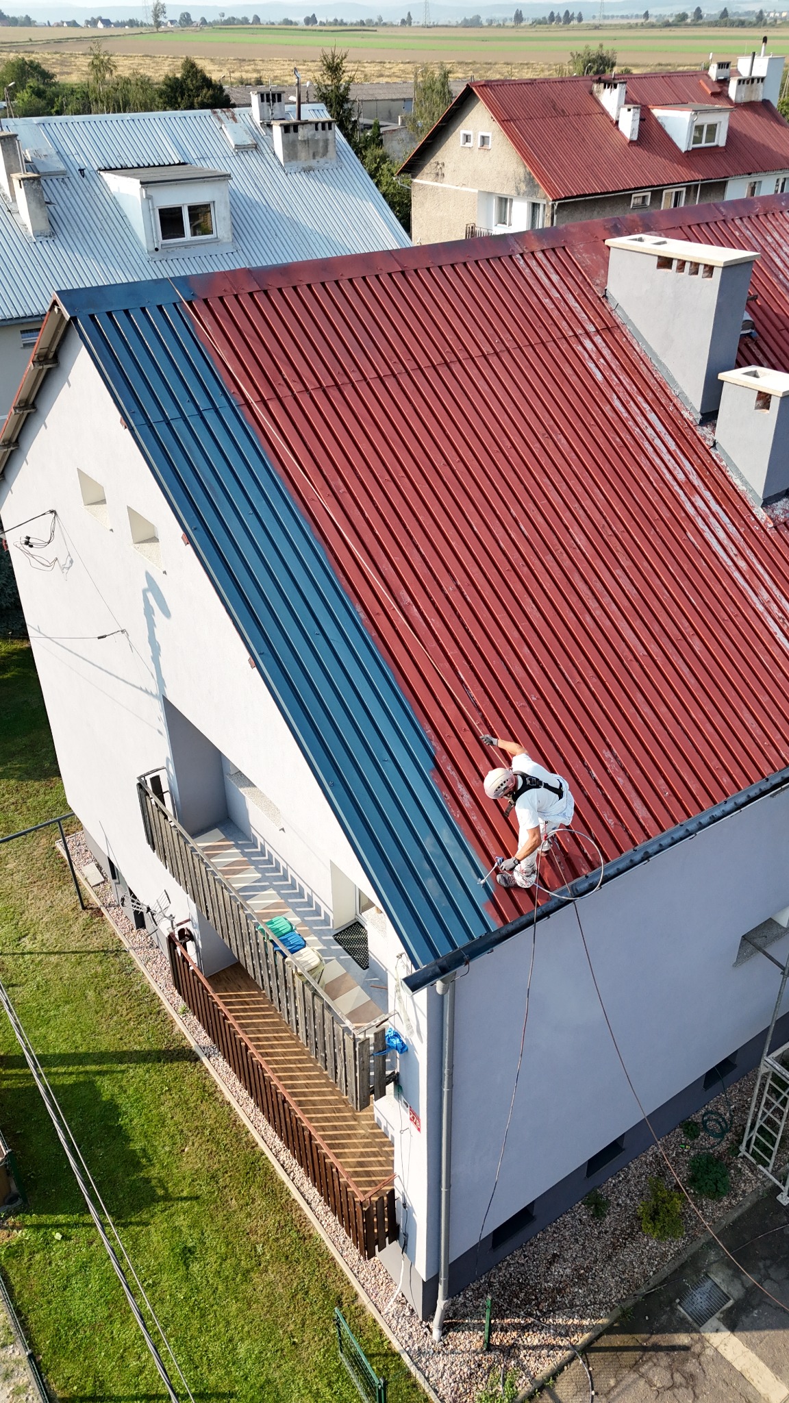Pracownik w uprzęży maluje blachodachówkę na czerwono, część dachu pomalowana na niebiesko, widok z góry na dom z balkonem.