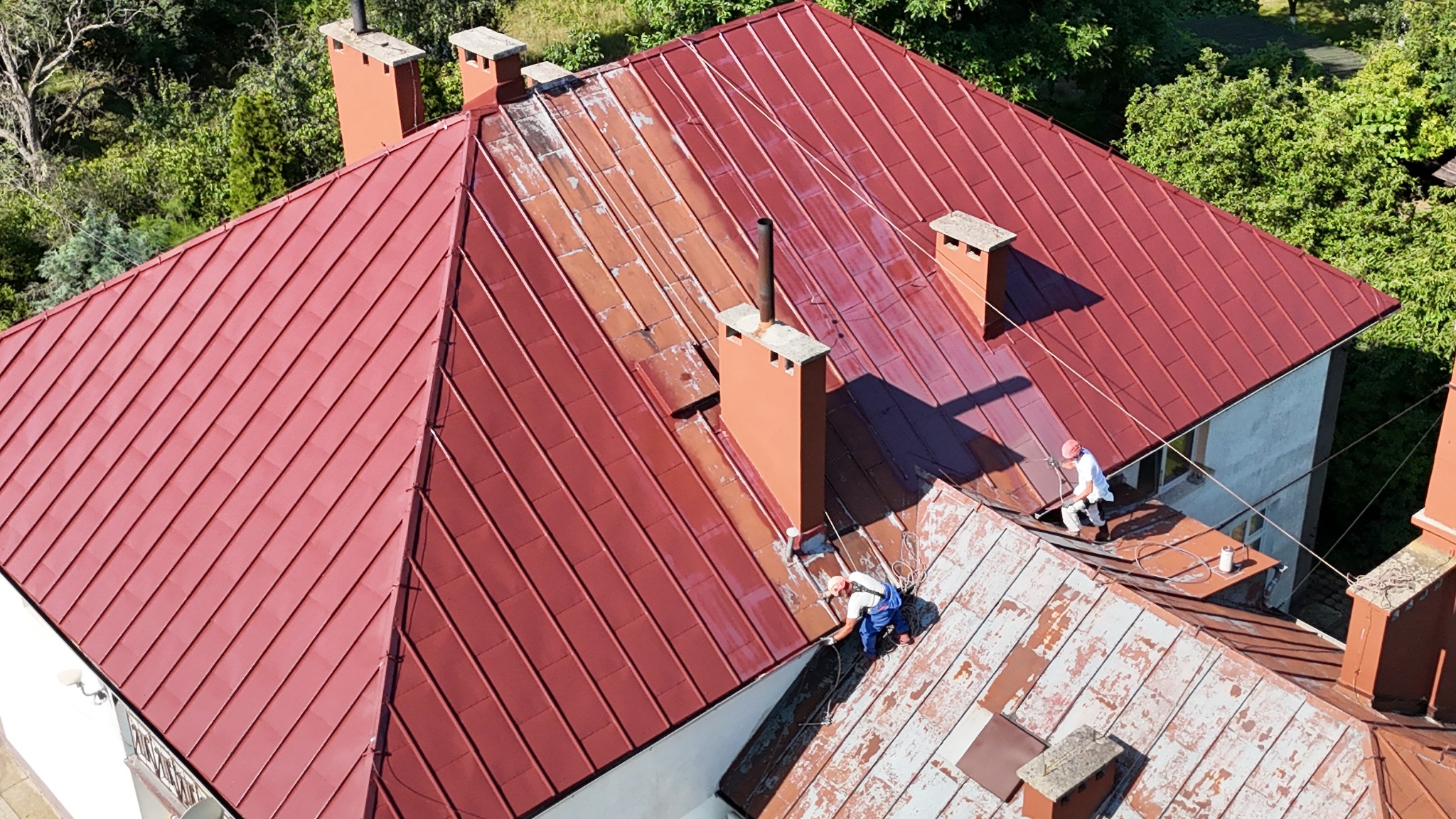 Częściowo umyty, czerwony dach z blachy trapezowej podczas pracy dwóch alpinistów na linach, widok z góry na dom z ceglanymi kominami i zielenią w tle.