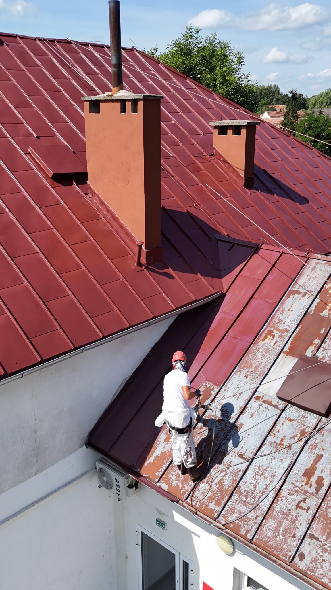 Pracownik w białym kombinezonie i czerwonym kasku maluje dach z blachy trapezowej na kolor ceglasty, widoczne fragmenty dachu z rdzawym nalotem, kominy w kolorze dachu, budynek z białą elewacją...