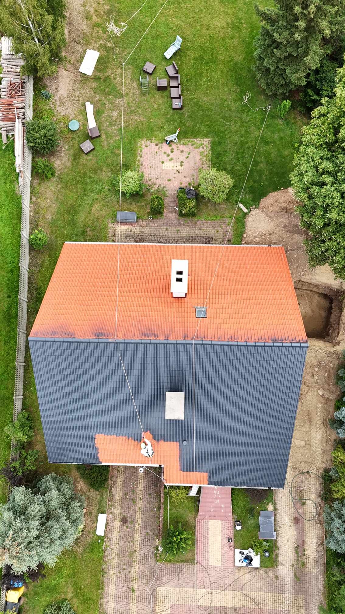 Widok z góry na dach domu, gdzie robotnik na linach zabezpieczających maluje fragment dachu na pomarańczowo, kontrastując z szarym, odnowionym fragmentem; na trawniku widoczne meble ogrodowe...