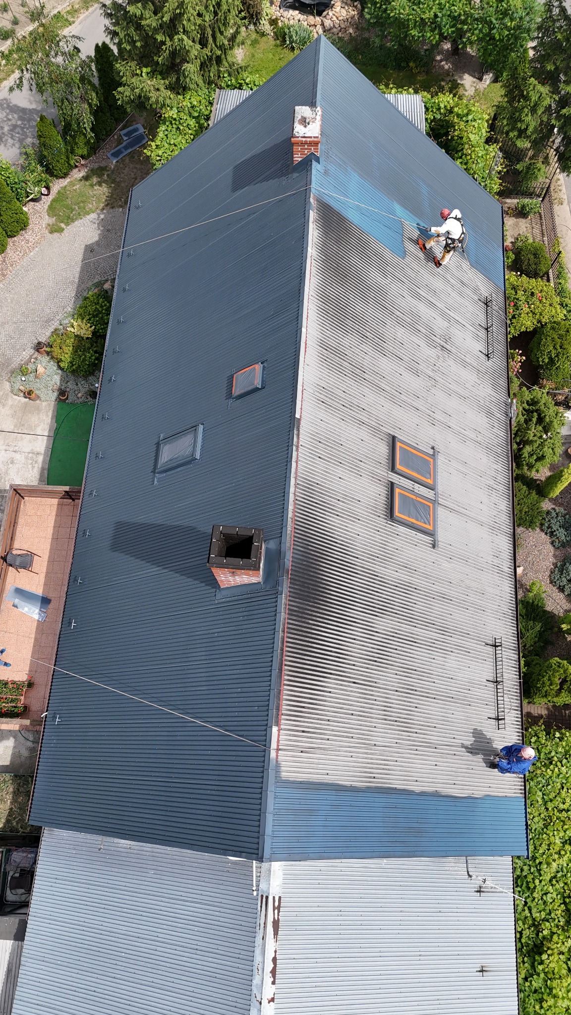 Widok z góry na dwuspadowy dach z blachy trapezowej, częściowo pomalowany na ciemnoszaro, z dwoma pracownikami na wysokościach, jeden w trakcie malowania, drugi w niebieskim kombinezonie przy...