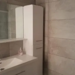 Wykończona łazienka z szarymi płytkami imitującymi kamień, biała szafka pod umywalkę z dwiema szufladami i wysoką szafką z lustrem, widoczna butelka mydła w płynie.