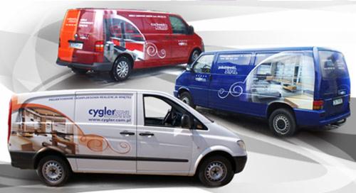 Trzy samochody dostawcze w różnych kolorach (biały, czerwony, niebieski) z grafikami przedstawiającymi wnętrza pomieszczeń i logo firm, zaparkowane na jasnym tle.