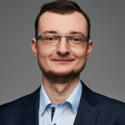 Portret uśmiechniętego mężczyzny w okularach i granatowej marynarce na szarym tle. Widoczna delikatna broda i starannie uczesane włosy.