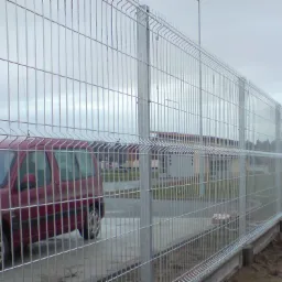 Długi, srebrny płot z paneli drucianych na betonowej podmurówce, widziany z perspektywy ulicy, z częściowo zasłoniętym czerwonym samochodem po lewej stronie.