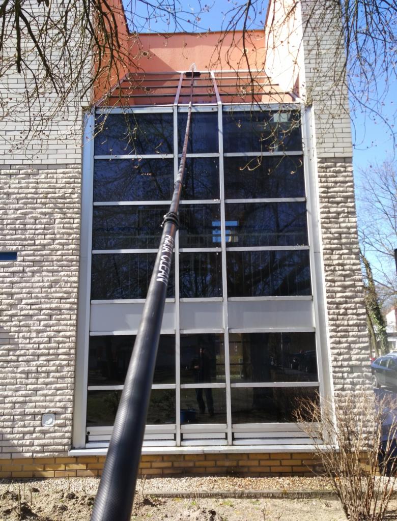 Czarny, teleskopowy kij z napisem 'CLEAN' sięga do wysokich okien budynku z cegły i betonu, odbijających drzewa i niebo.