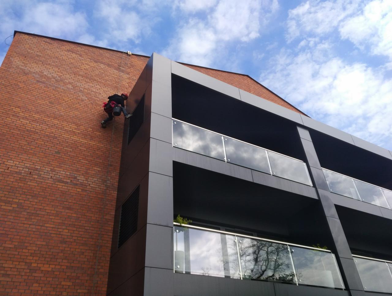 Pracownik w kasku, na linie, myje okna na wysokości w Szczodrzykowie. Widok z dołu na ceglany budynek z balkonami.