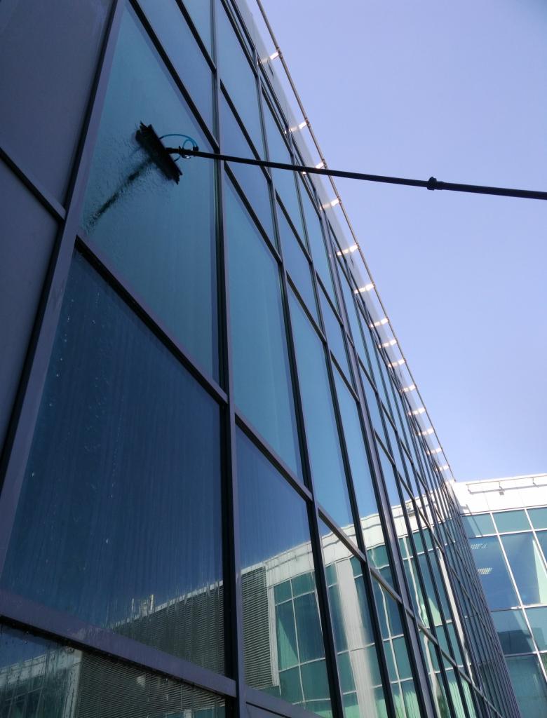 Czyszczenie szklanej fasady biurowca za pomocą teleskopowej lancy z szczotką i dopływem wody, widok z dołu na tle błękitnego nieba.