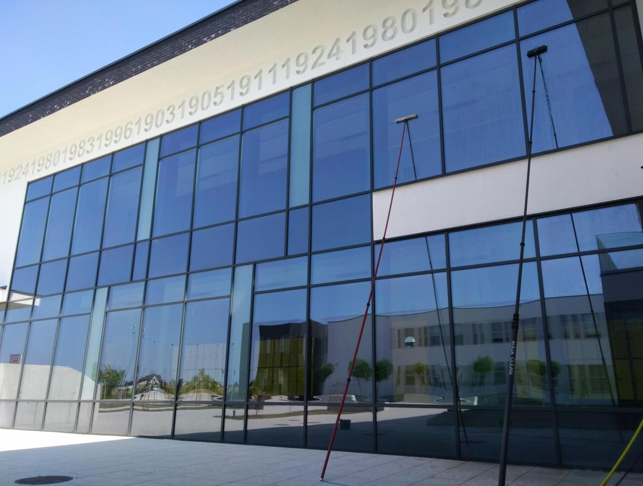 Nowoczesny budynek biurowy z dużą ilością okien, czyszczony za pomocą długich tyczek z myjkami; na elewacji widoczne numery.