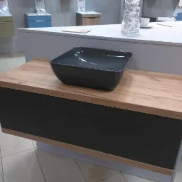 Ciemnoszara, kwadratowa umywalka nablatowa na szafce z drewnopodobnym blatem i czarnymi frontami, ekspozycja w salonie łazienek.