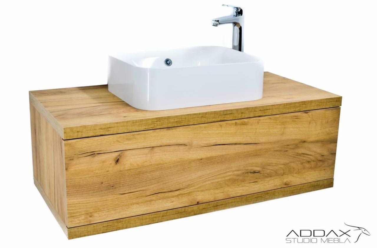 Szafka łazienkowa z blatem z drewna dębowego i białą, kwadratową umywalką nablatową oraz chromowaną baterią, na białym tle, z widocznym logo producenta w prawym dolnym rogu.