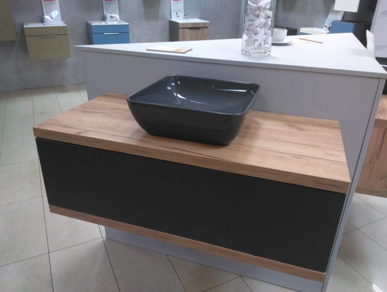 Ciemnoszara, kwadratowa umywalka nablatowa na szafce z drewnopodobnym blatem i czarnymi frontami, ekspozycja w salonie łazienek.