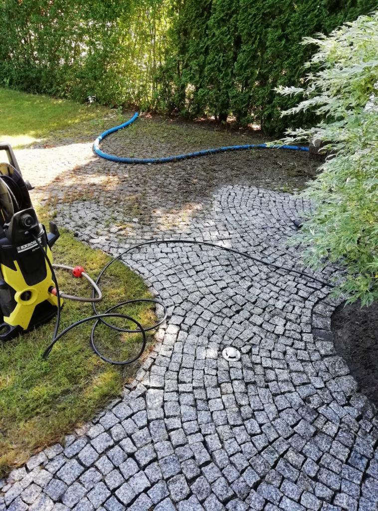 Umyta kostka brukowa na ścieżce ogrodowej, widoczny wąż ciśnieniowy i myjka ciśnieniowa Karcher, trawnik i roślinność w tle.