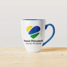 Biały kubek z niebieskim wnętrzem i uchwytem, nadrukowane logo Powiatu Pińczowskiego z elementami w kolorach żółtym, niebieskim i zielonym, stojący na jasnym drewnianym blacie.