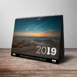Kalendarz biurkowy 2019 z pejzażem o wschodzie słońca, leżący na drewnianym blacie, z widocznym adresem strony internetowej.