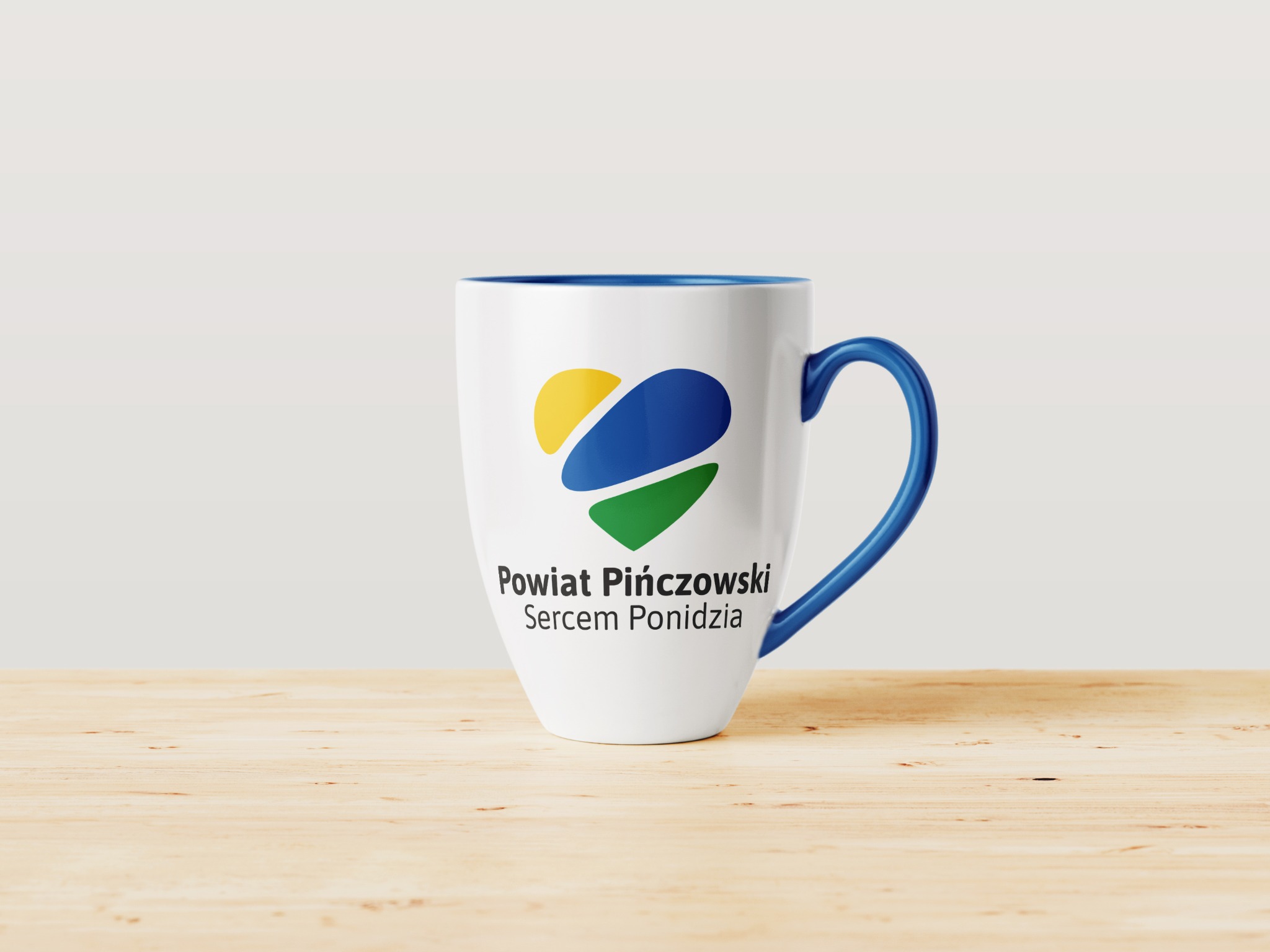Biały kubek z niebieskim wnętrzem i uchwytem, nadrukowane logo Powiatu Pińczowskiego z elementami w kolorach żółtym, niebieskim i zielonym, stojący na jasnym drewnianym blacie.