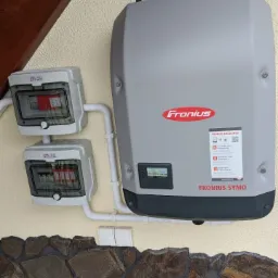 Szary inwerter Fronius Symo zamontowany na elewacji budynku obok dwóch skrzynek bezpiecznikowych ETI, widoczne fragmenty instalacji elektrycznej i kamiennej podmurówki.