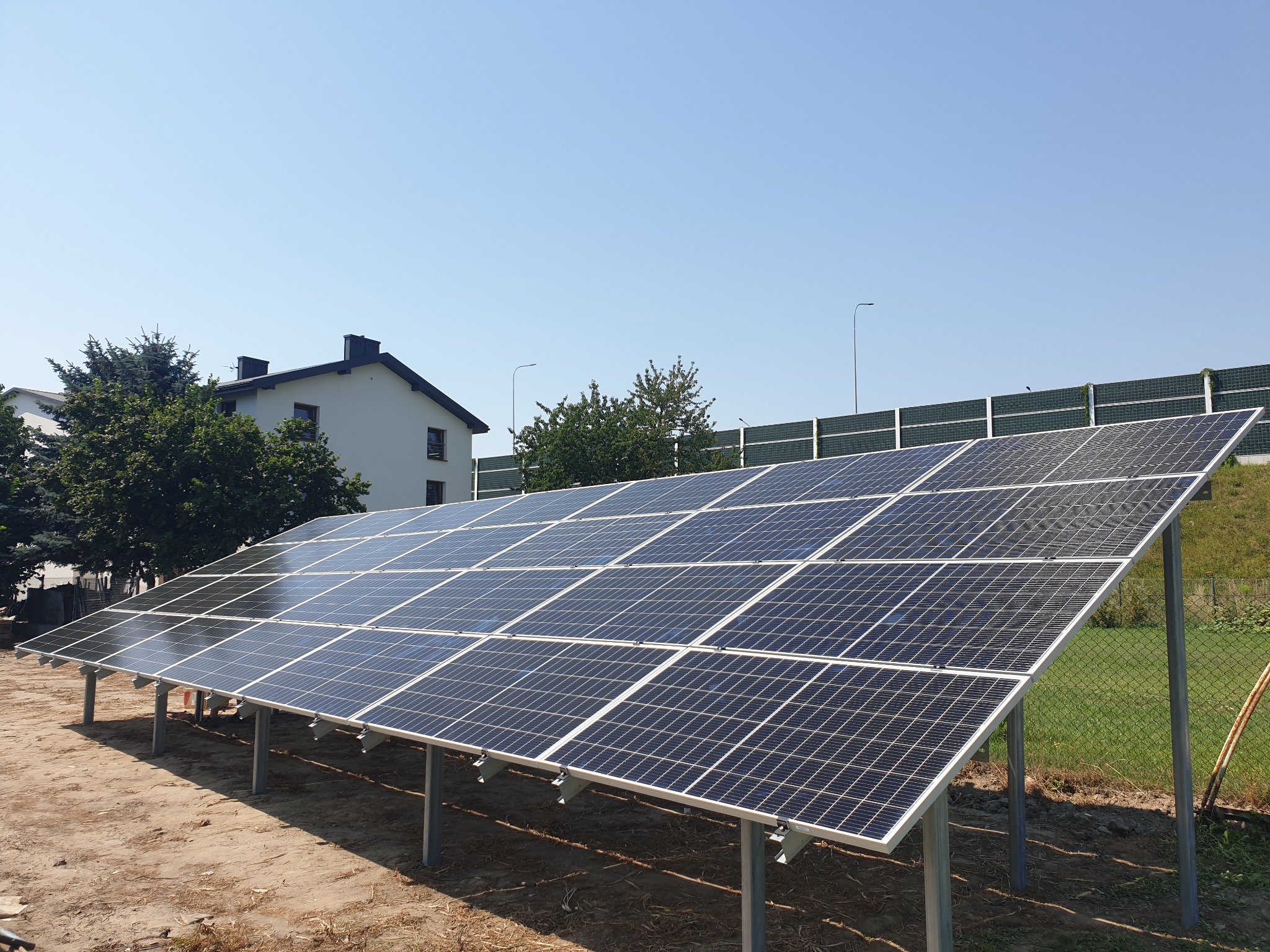 Instalacja paneli słonecznych na konstrukcji wsporczej, widoczny budynek mieszkalny w tle i barierki dźwiękochłonne.