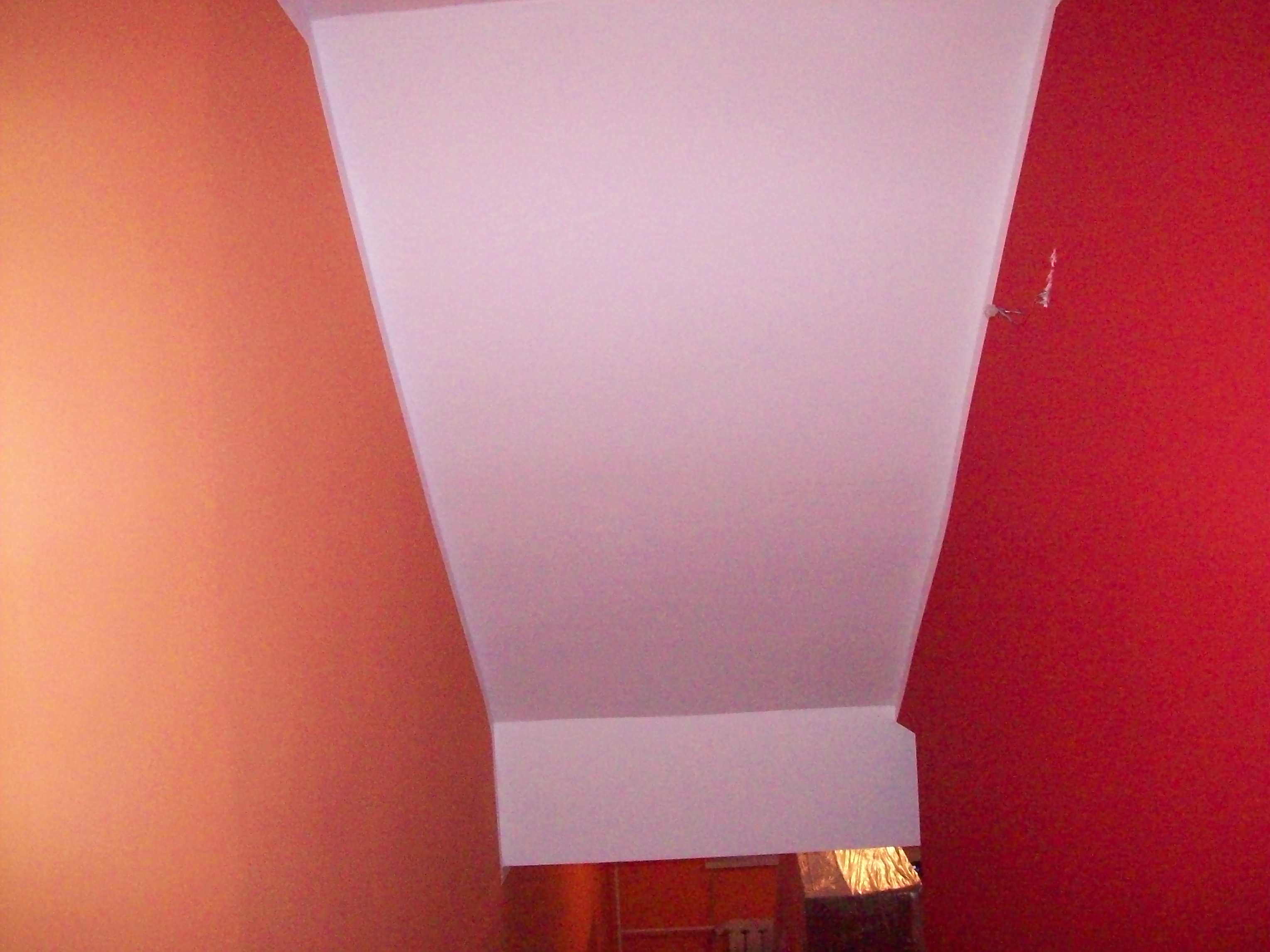 Pomarańczowa i czerwona ściana w pomieszczeniu z widocznym fragmentem białego sufitu z narożnikiem, oraz fragmentem grzejnika i lampy w tle.