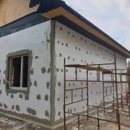 Dom w trakcie ocieplania styropianem, z widocznym rusztowaniem i fragmentem dachu pokrytego papą. Ślady po kleju na styropianie przed nałożeniem siatki.