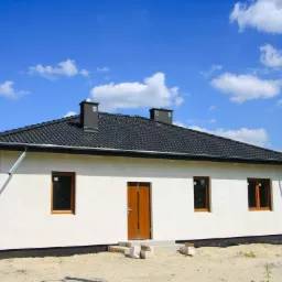 Nowo wybudowany parterowy dom z ciemną dachówką, beżową elewacją i brązowymi ramami okiennymi, widoczny fragment rynny i komin, otoczony piaszczystym terenem.