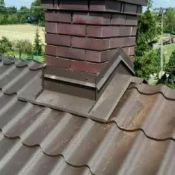 Brązowy dach z blachodachówki z nowo wymurowanym kominem z cegły klinkierowej i obróbką blacharską, widok na tle zieleni i błękitnego nieba.