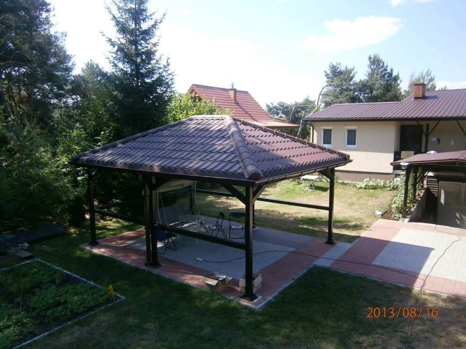 Zadaszona altana z brązową dachówką na betonowej wylewce obramowanej czerwoną kostką, widok z góry na tle domu i ogrodu.