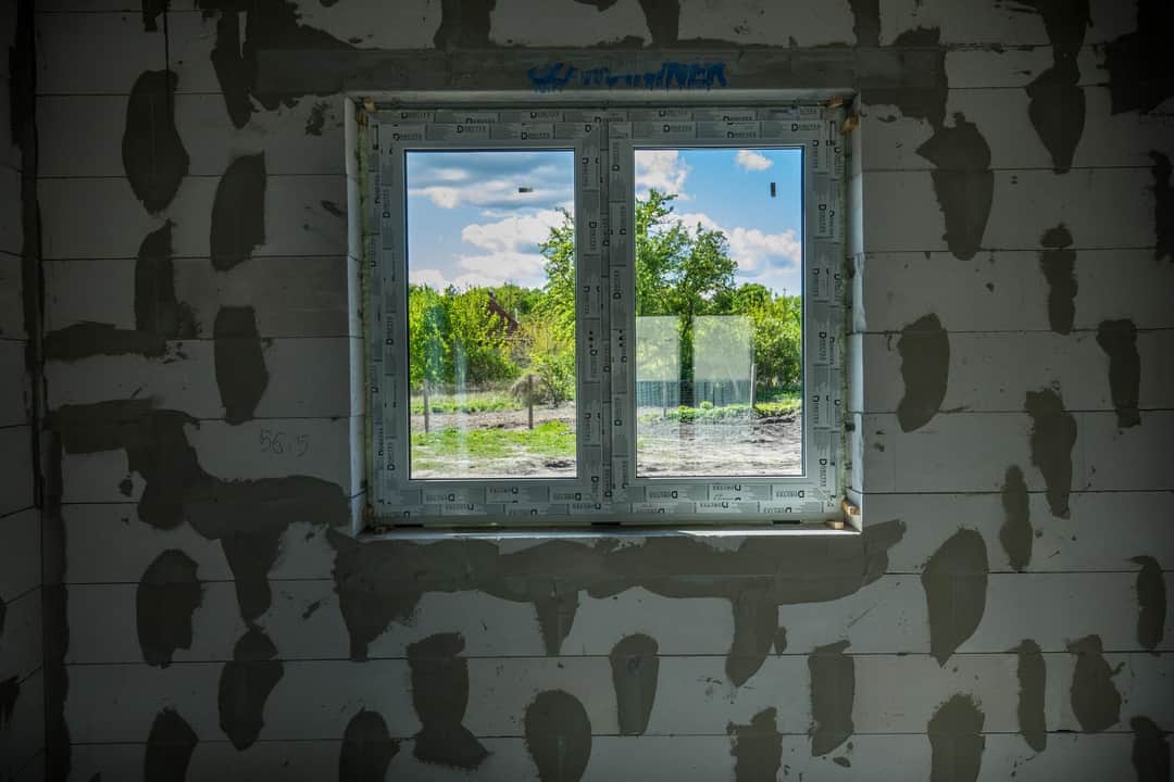 Dwuszybowe okno PCV zamontowane w surowej ścianie z betonu komórkowego, widok na zewnątrz z zielenią i niebem.