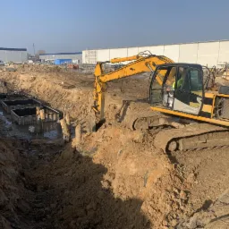 Żółta koparka gąsienicowa pracuje przy wykopie pod fundamenty z szalunkami, widoczne zbrojenia, w tle hala magazynowa i błękitne niebo.