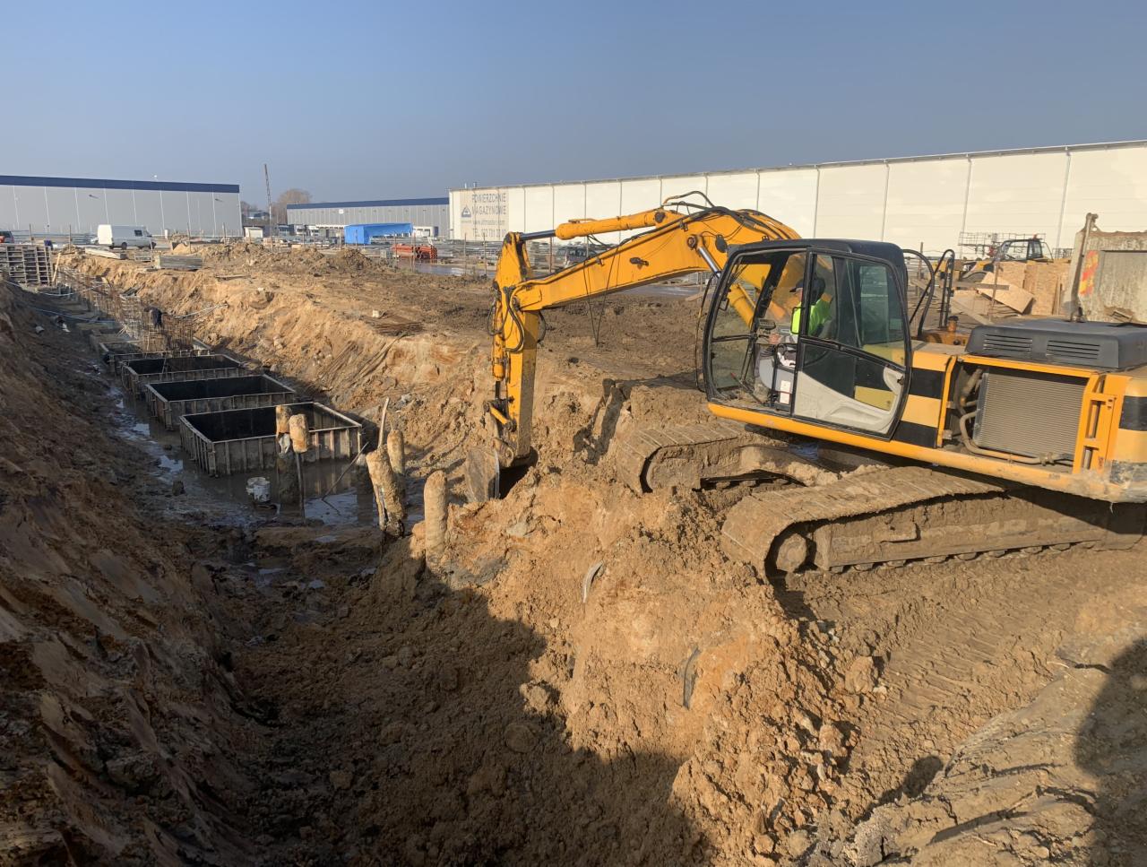 Żółta koparka gąsienicowa pracuje przy wykopie pod fundamenty z szalunkami, widoczne zbrojenia, w tle hala magazynowa i błękitne niebo.