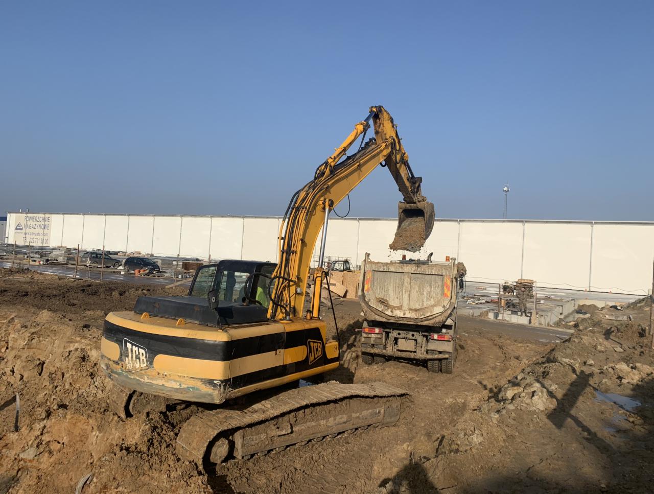 Żółto-czarna koparka JCB nabiera ziemię i wrzuca ją do brudnej wywrotki na placu budowy, w tle biały magazyn.