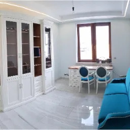 Jasne wnętrze gabinetu z białą zabudową biblioteczną, biurkiem z krzesłami i turkusową sofą na marmurowej podłodze.